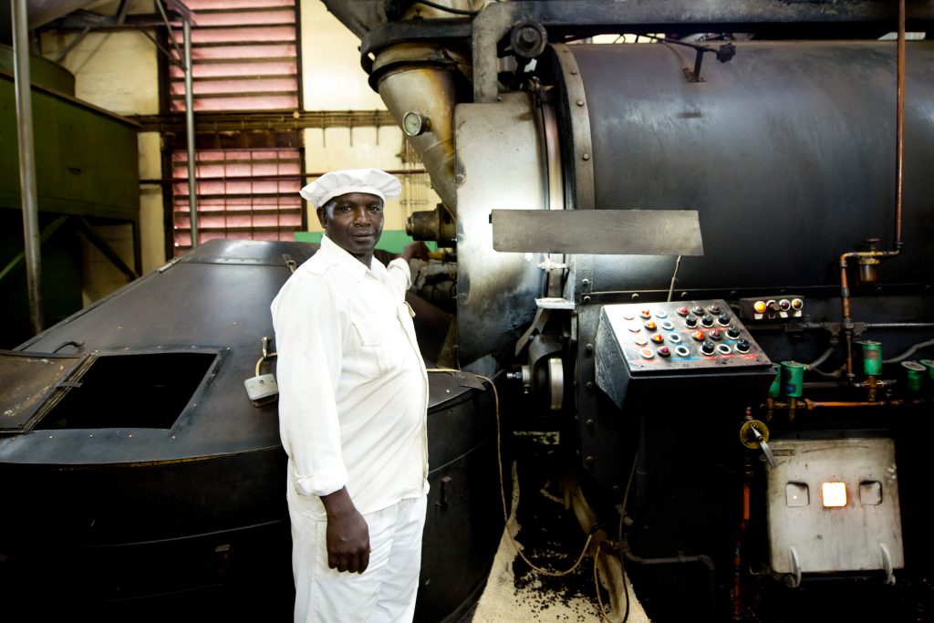Das Foto zeigt einen Mitarbeiter im Vordergrund. Im Hintergrund sehen Sie die Kaffeeröstung. Das Foto ist bei der Kaffee Reise beim Besuch der Bio Fairtrade Kaffee und der Betriebsbesichtigung der Bio Fairtrade Kaffee Kooperative Bauern in Tansania entstanden. Wir führen durch alle Schritte der Kaffee Herstellung. Sie sehen das Rösten der Kaffeebohnen.
